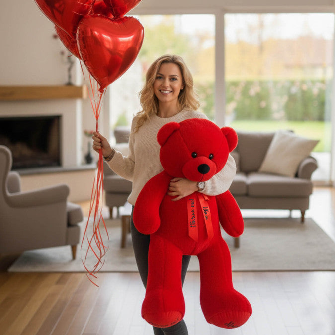Woman holding a large red teddy bear and heart-shaped balloons in a living room.
