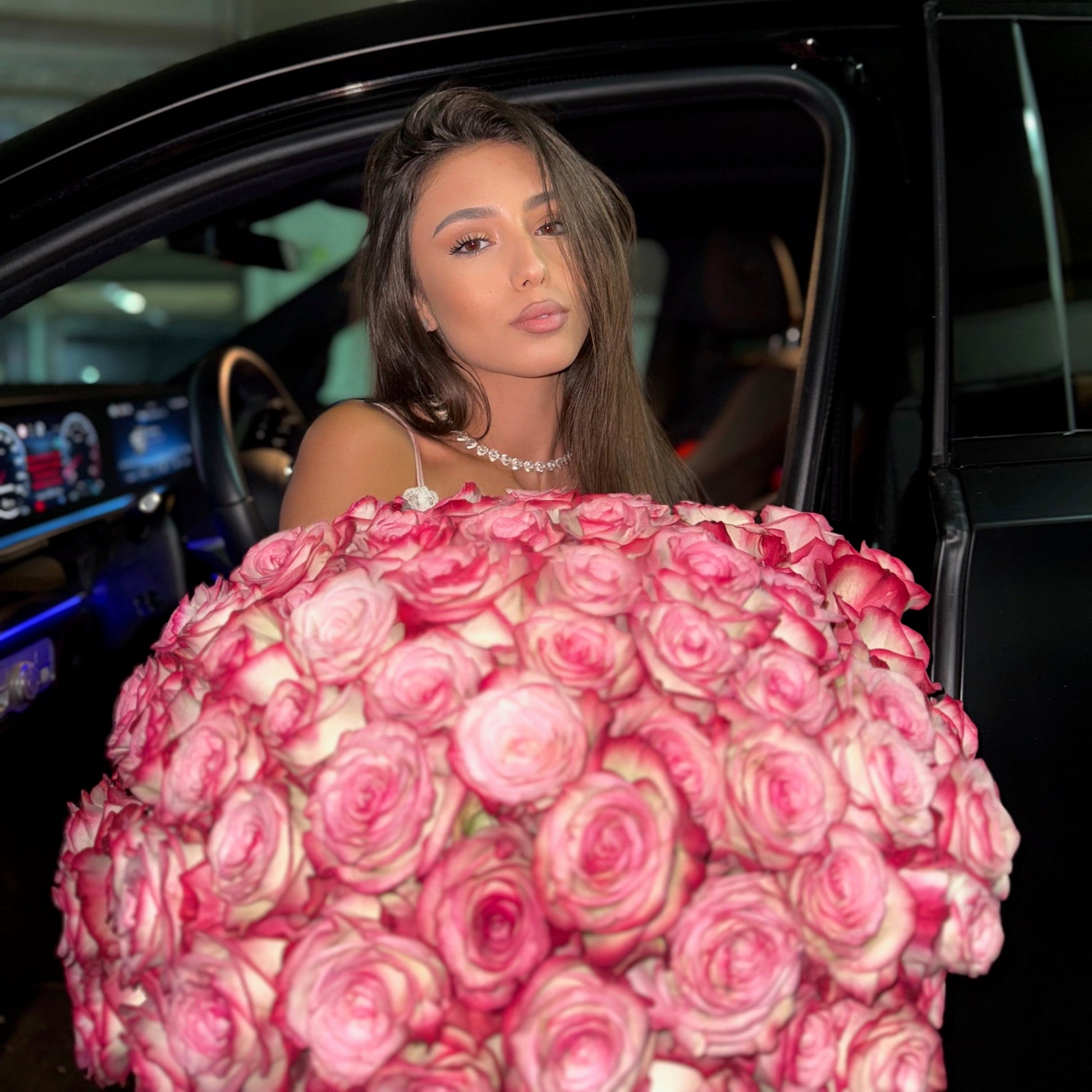 Woman holding a large bouquet of pink roses in front of a car.