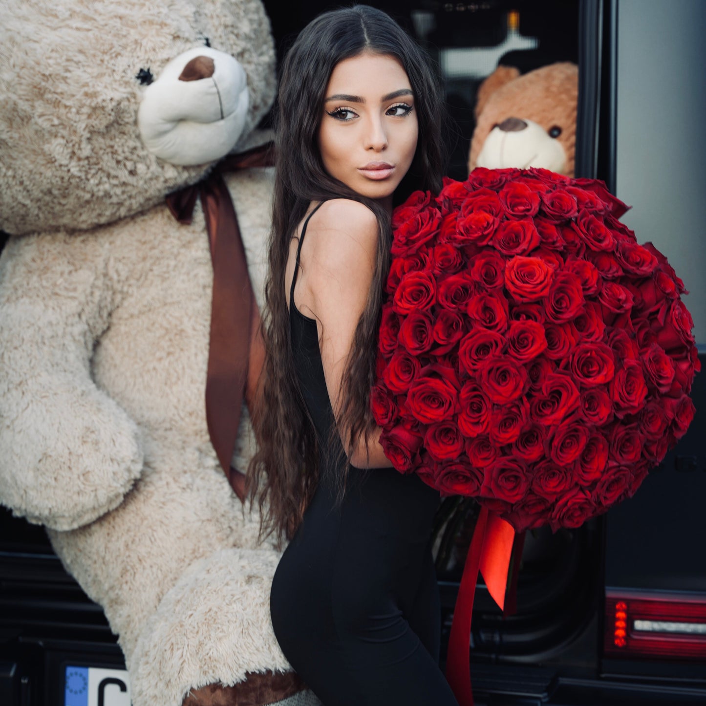 Woman holding a large bouquet of red roses next to a giant teddy bear in a car.