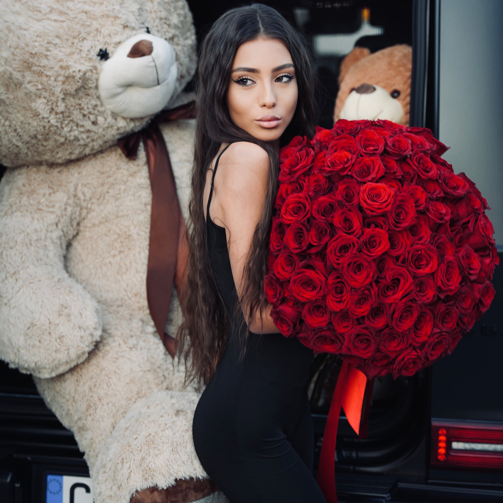 Woman holding a large bouquet of red roses next to a giant teddy bear in a car.
