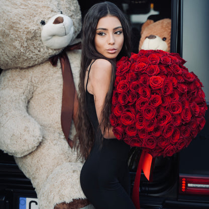 Woman holding a large bouquet of red roses next to a giant teddy bear in a car.