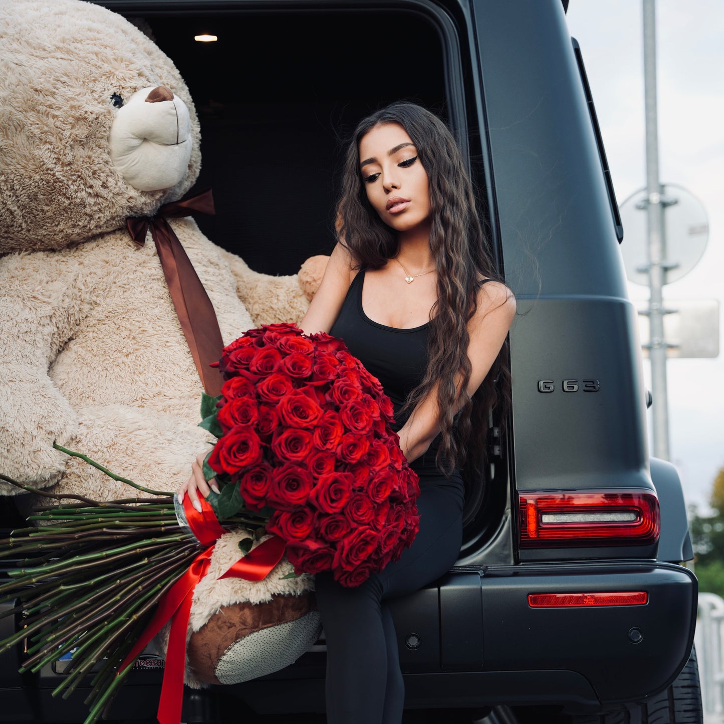 Woman sitting in a car with a large teddy bear and bouquet of red roses.