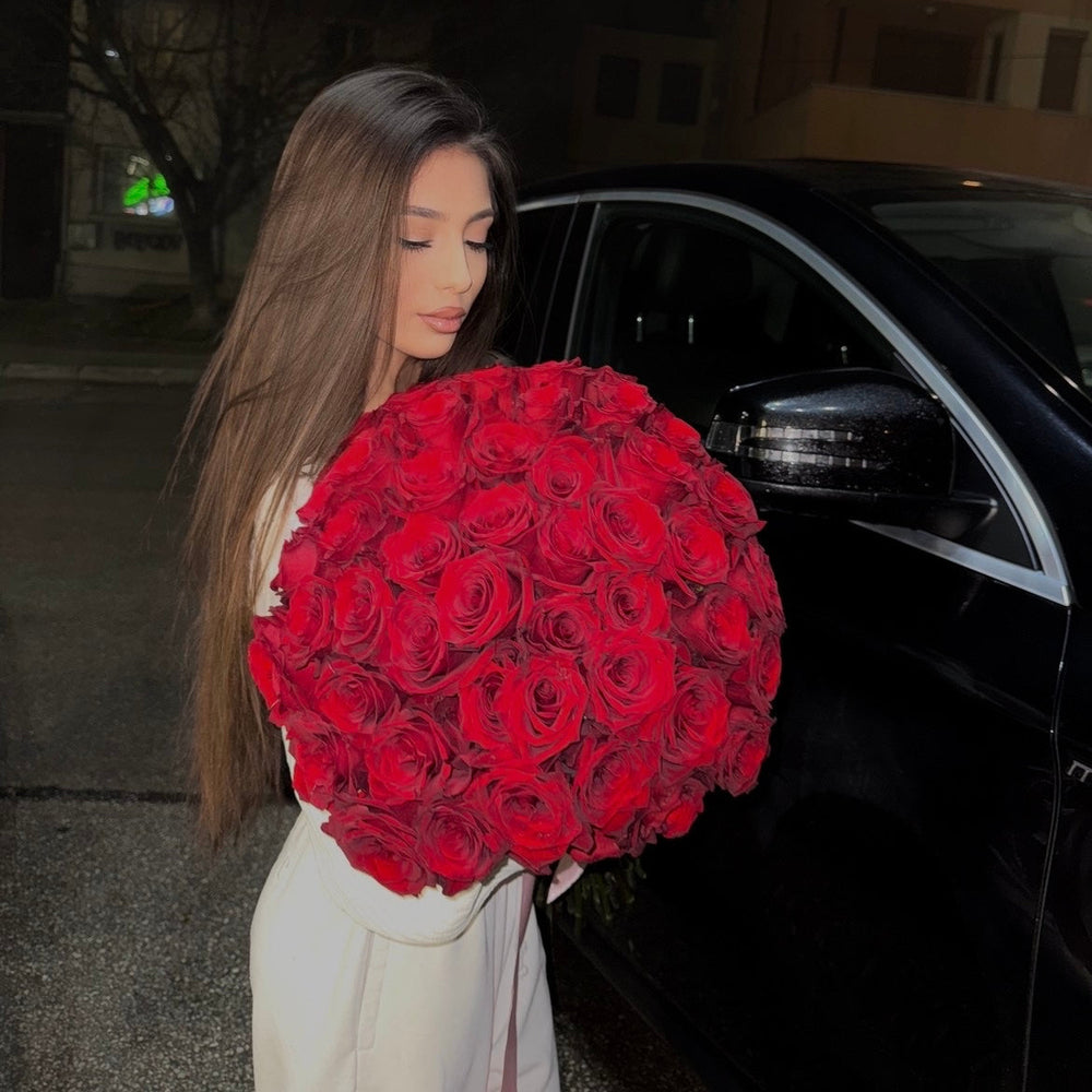 Woman holding a large bouquet of red roses in front of a black car.