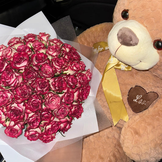 Teddy bear with a gold bow and bouquet of red roses on a reflective surface