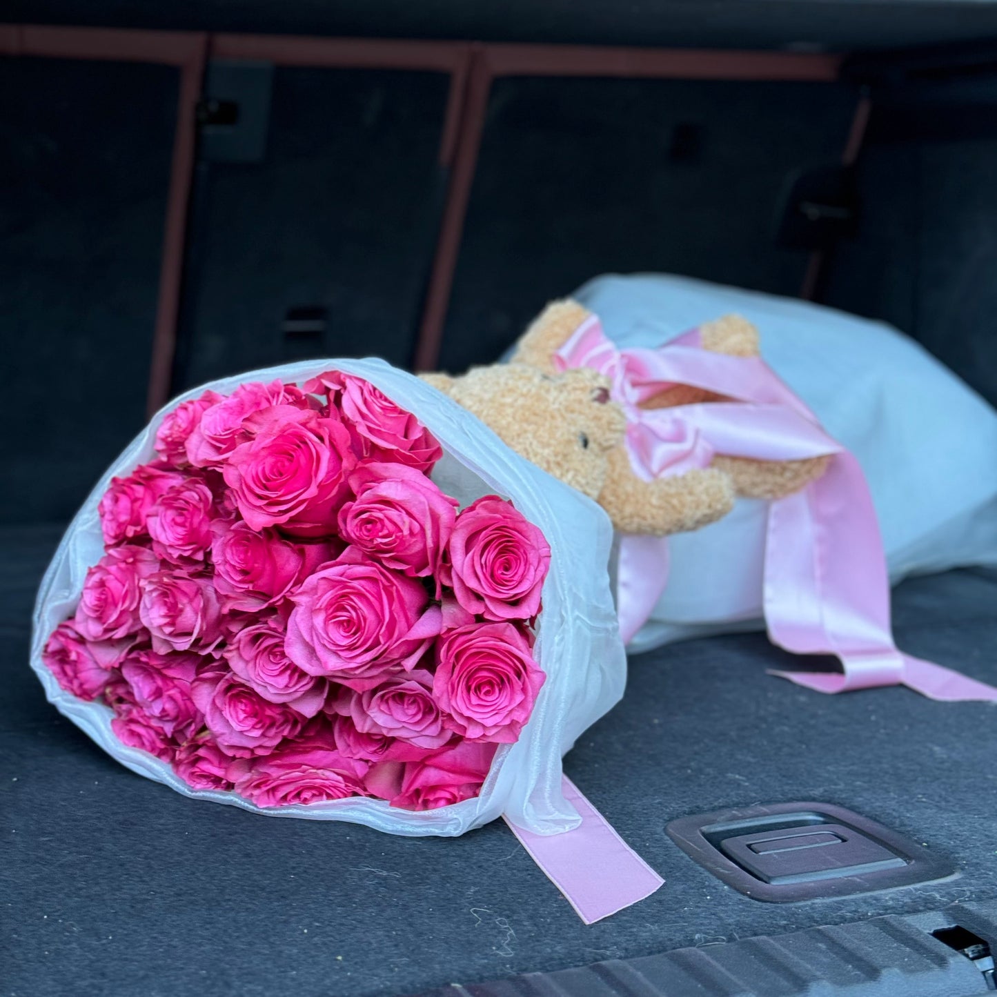 Bouquet of pink roses and teddy bear with pink ribbon in a car trunk.