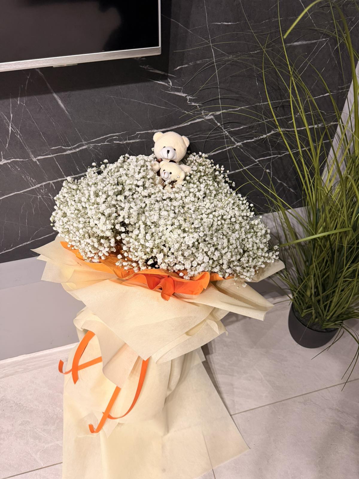 Bouquet of baby's breath with teddy bears on top, wrapped in orange ribbon, against a marble wall.