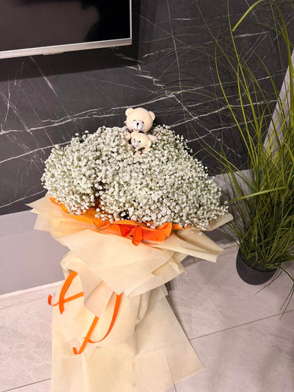Bouquet of baby's breath with teddy bears on top, wrapped in orange ribbon, against a marble wall.