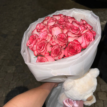 Bouquet of pink roses held by a hand with a white teddy bear wearing a pink bow tie.
