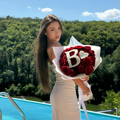 Woman holding a bouquet of red roses with 'B' letters by a pool with greenery in the background. 